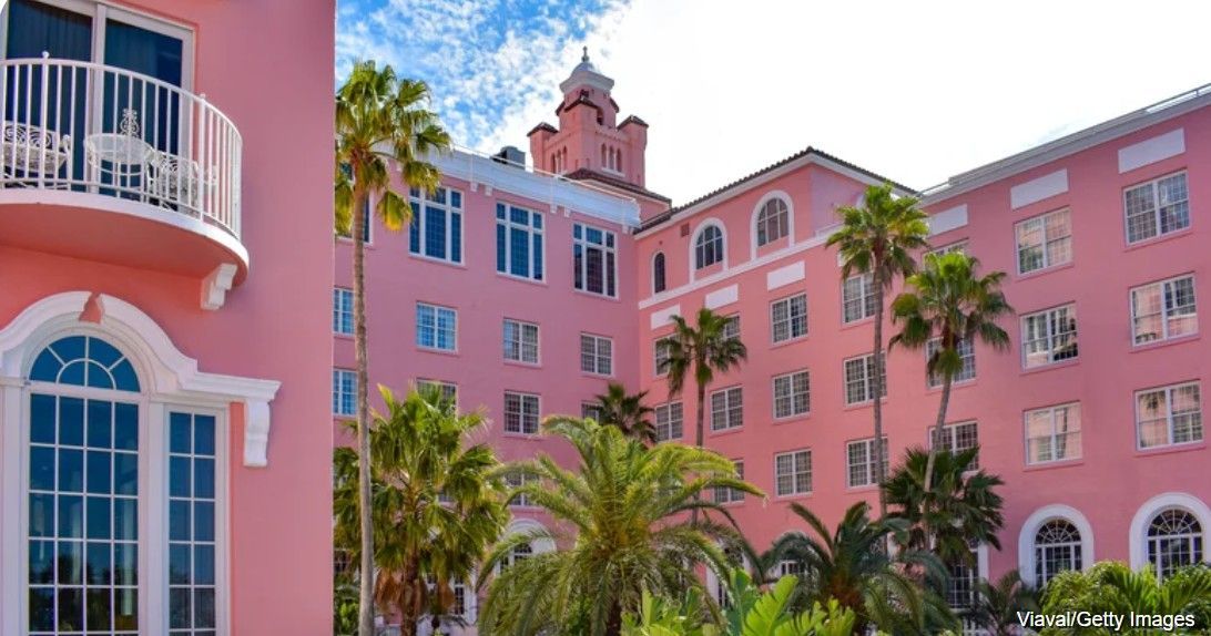 Pink resort building with palm trees and a balcony on a sunny day.