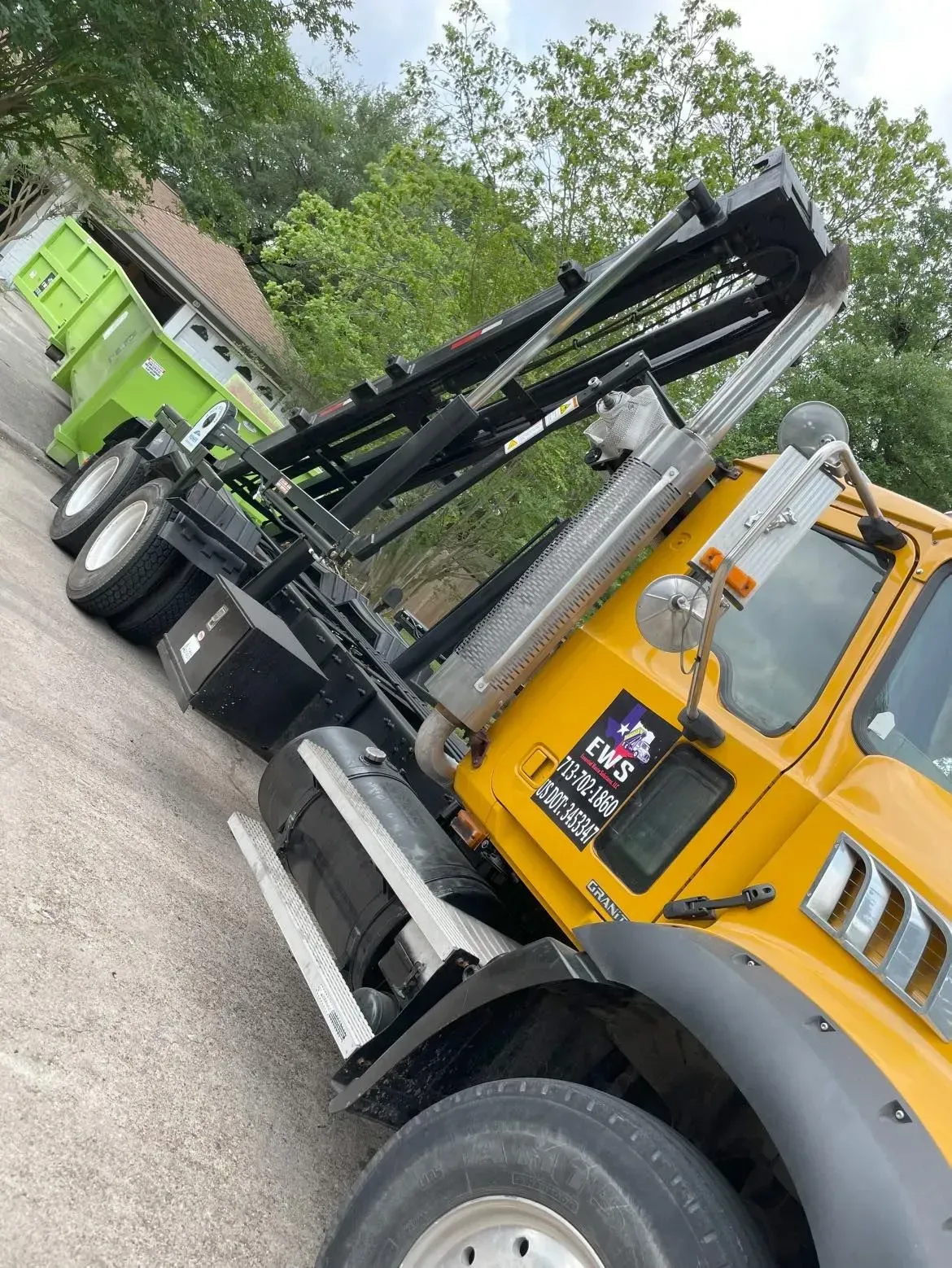 Yellow and black roll-off truck with a green dumpster in the background. Truck is raising the roll-off.
