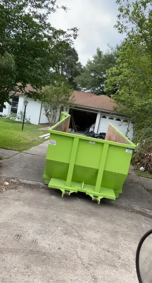 Green dumpster in a driveway in front of a white house with a partially open garage. Overcast sky.