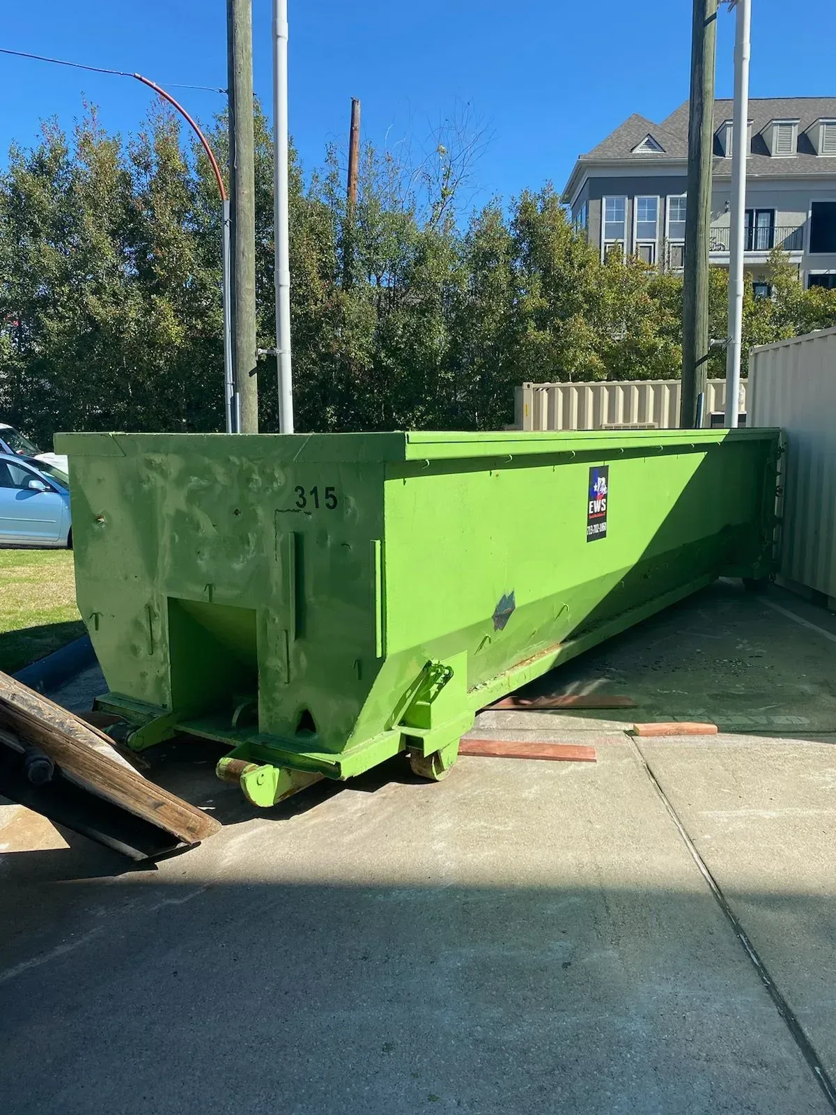 Green dumpster outside with a building and trees in the background.