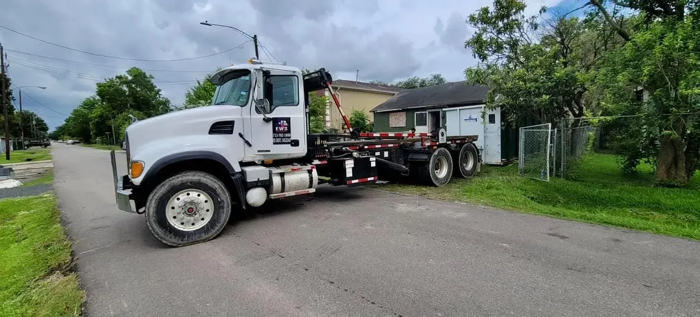 White truck with a hook lift parked on a road beside a small building. Overcast sky.