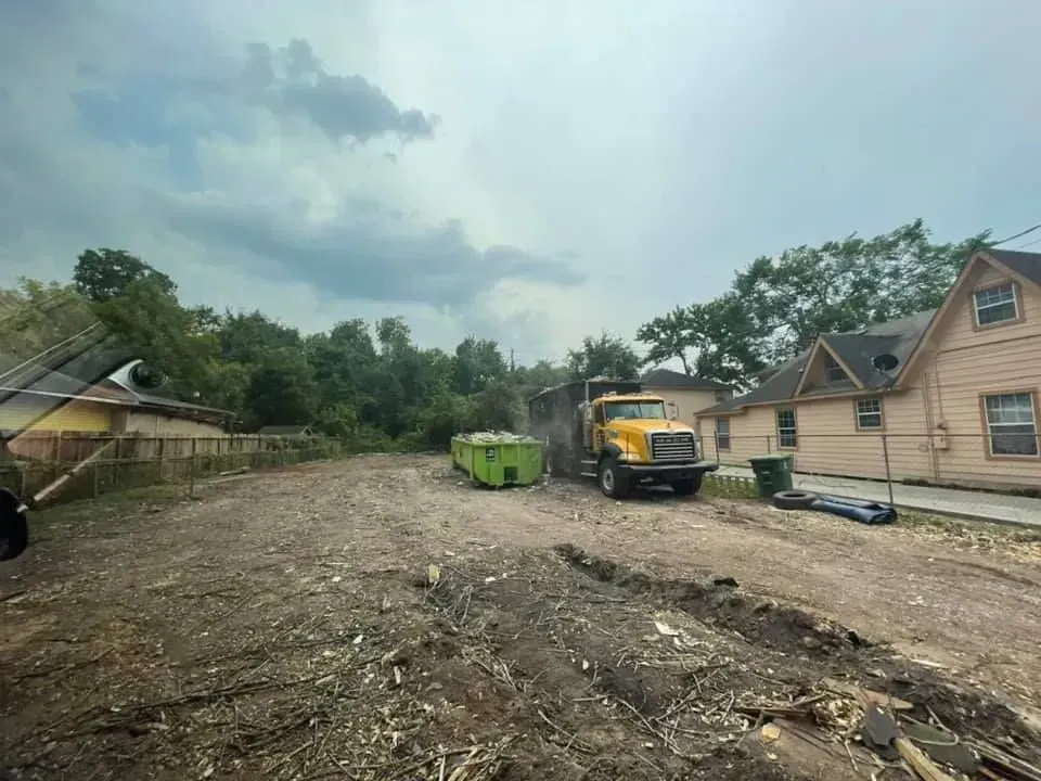 Dirt lot with yellow truck, green dumpster, and residential buildings on a cloudy day.