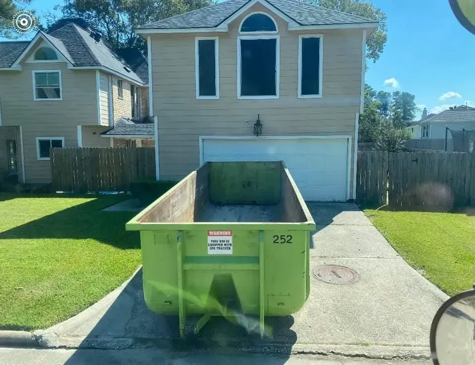 Green dumpster parked in front of a beige house with a driveway; sunny day.