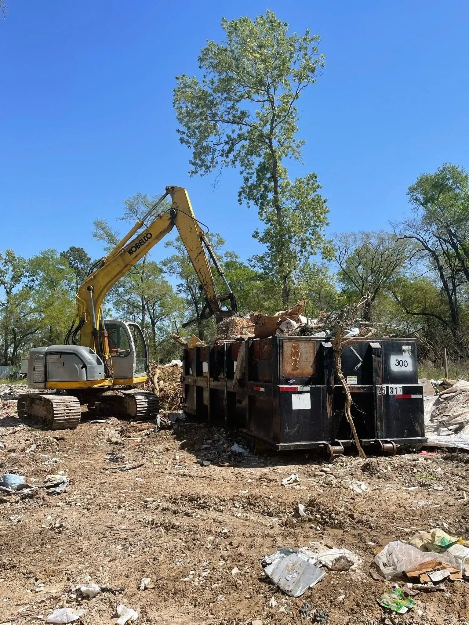 Excavator loading debris into a dumpster outdoors on a sunny day.