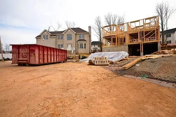 Construction site: new house under framing, brick home, red dumpster, dirt and gravel.