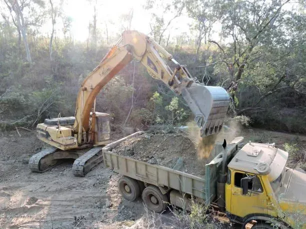 Yellow excavator loading a dump truck with dirt in a wooded area.