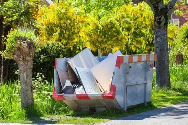 Metal dumpster overflowing with construction debris, beside a tree on a grassy verge, green trees in background.