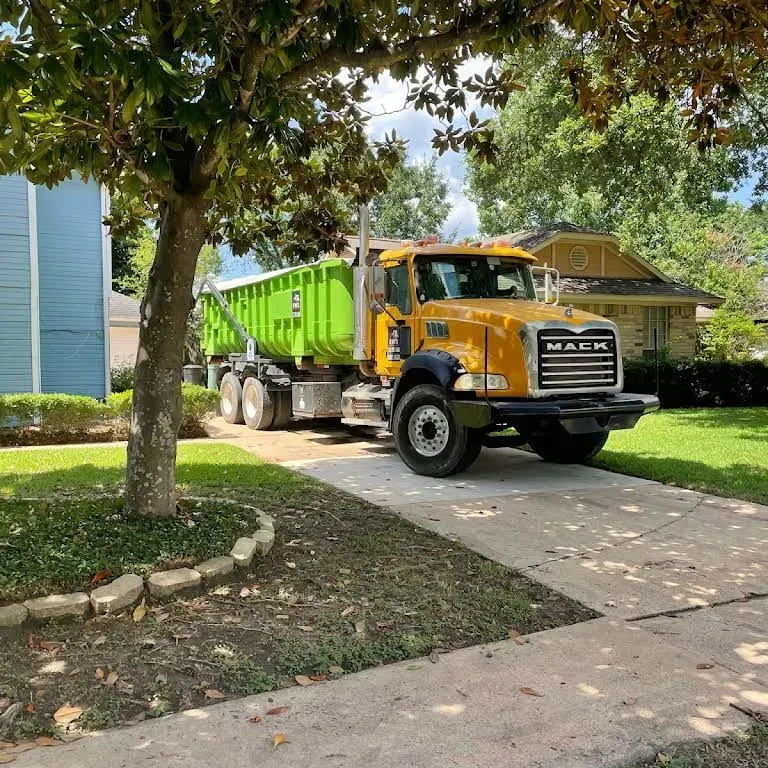 Yellow Mack dump truck parked on a residential driveway, lime green container bed, blue and yellow houses.