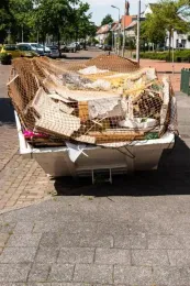 A white dumpster overflowing with debris, covered by a brown net, on a street.