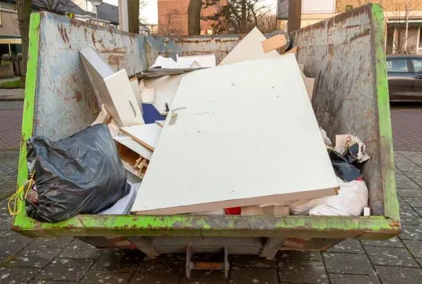 Green dumpster filled with debris including wood, a white door, and trash bags on a city street.