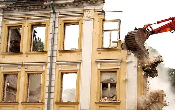 Demolition of a yellow building by an excavator, kicking up dust.