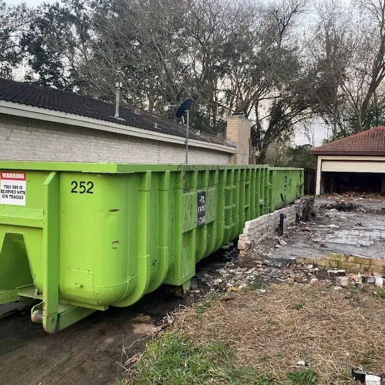 Large green dumpster beside a house undergoing demolition; debris, concrete blocks, and a garage are visible.