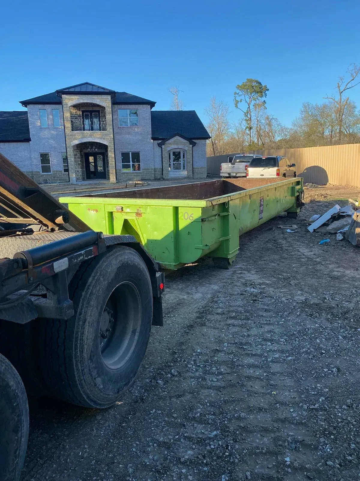 Bright green dumpster on a truck bed next to a large, unfinished house on a gravel lot.
