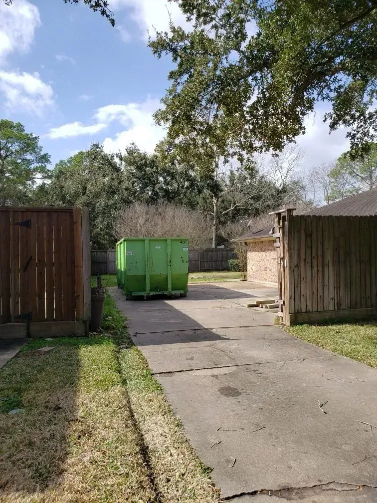 Green dumpster on a concrete driveway, flanked by wooden fences and grass, under a blue sky.