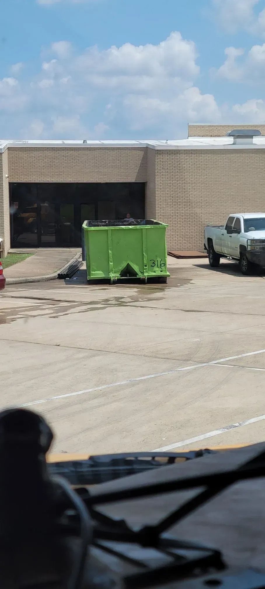 Green dumpster in front of a brick building. White truck parked nearby on a sunny day.