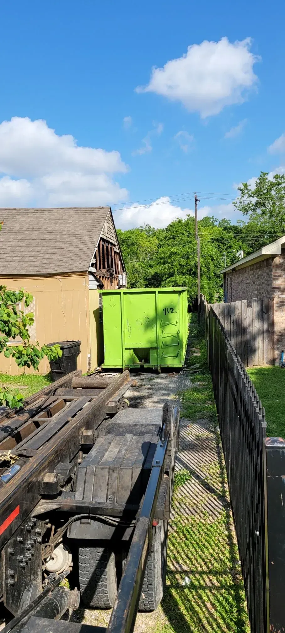 A green dumpster sits between buildings with a truck's mechanism in the foreground, under a blue sky.
