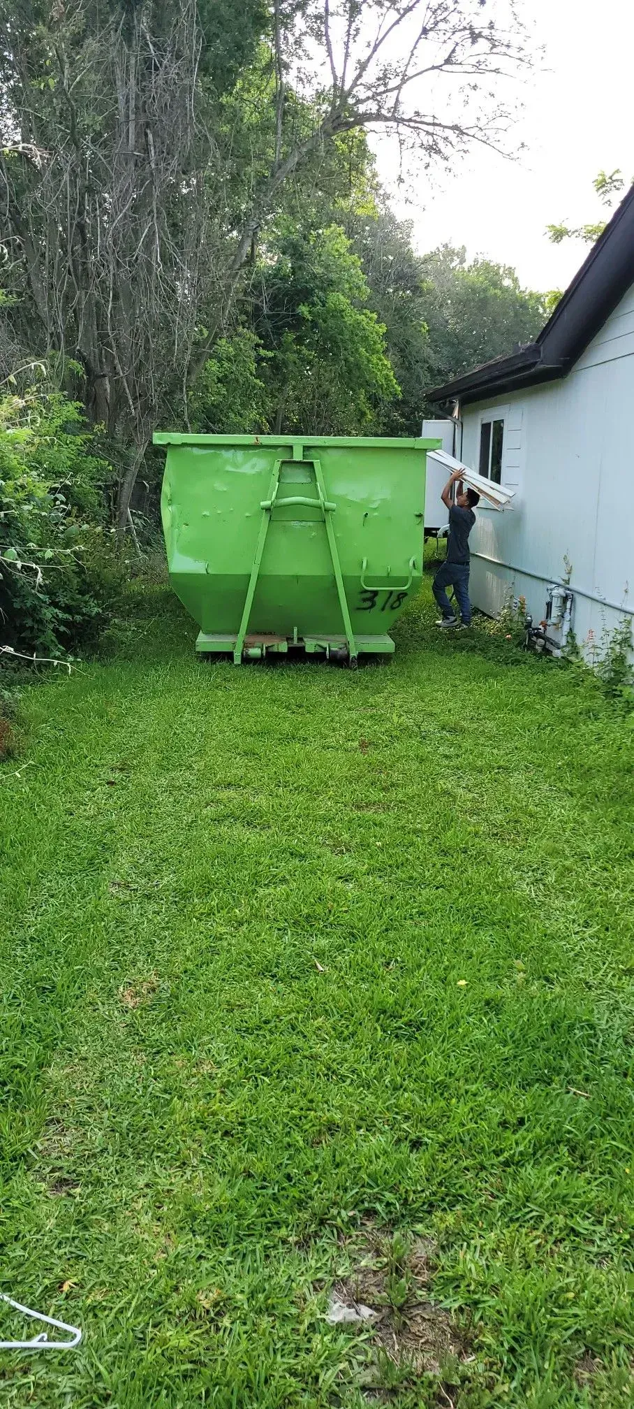 Green dumpster on a grassy lawn next to a building. A person stands near the building. Lush trees are in the background.