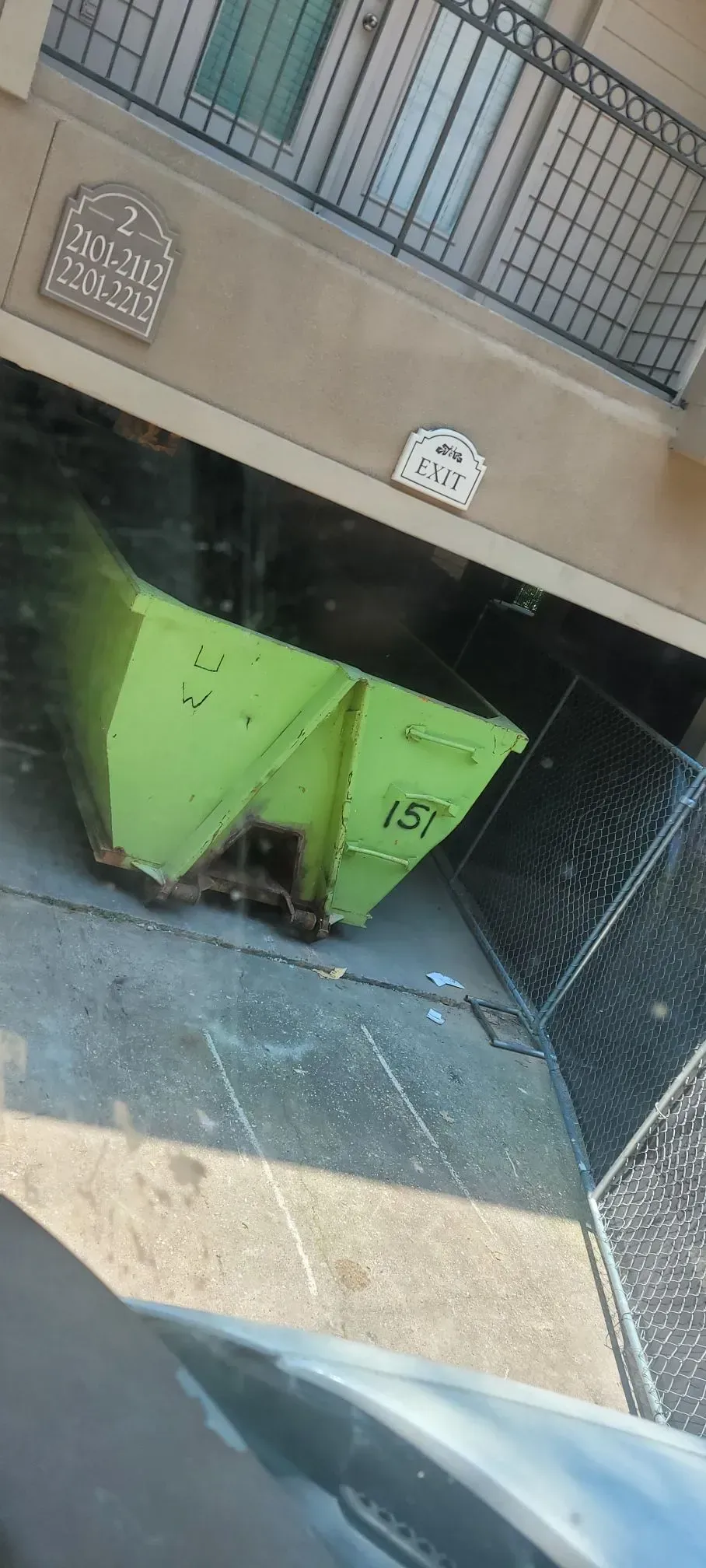 Green dumpster sits on a concrete pad near a building with wrought iron railings.