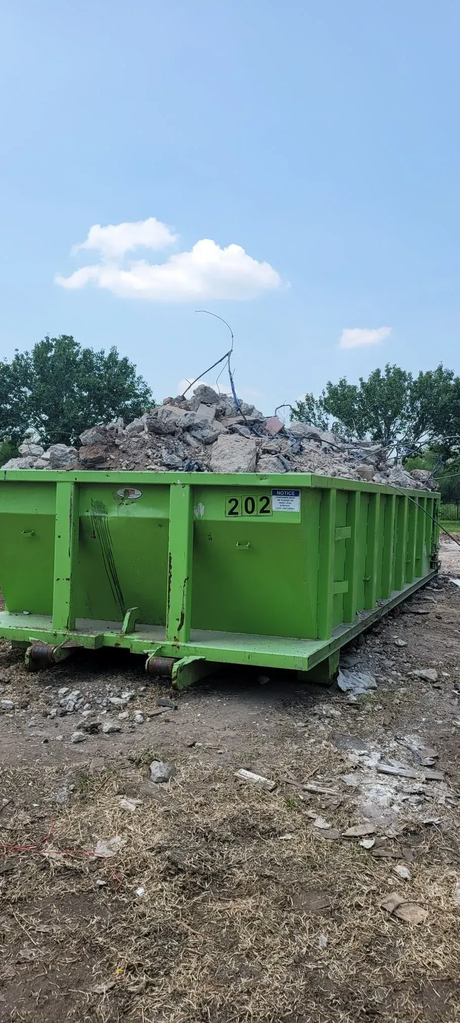 Green dumpster filled with rubble on dirt, under a blue sky. Trees in the background.