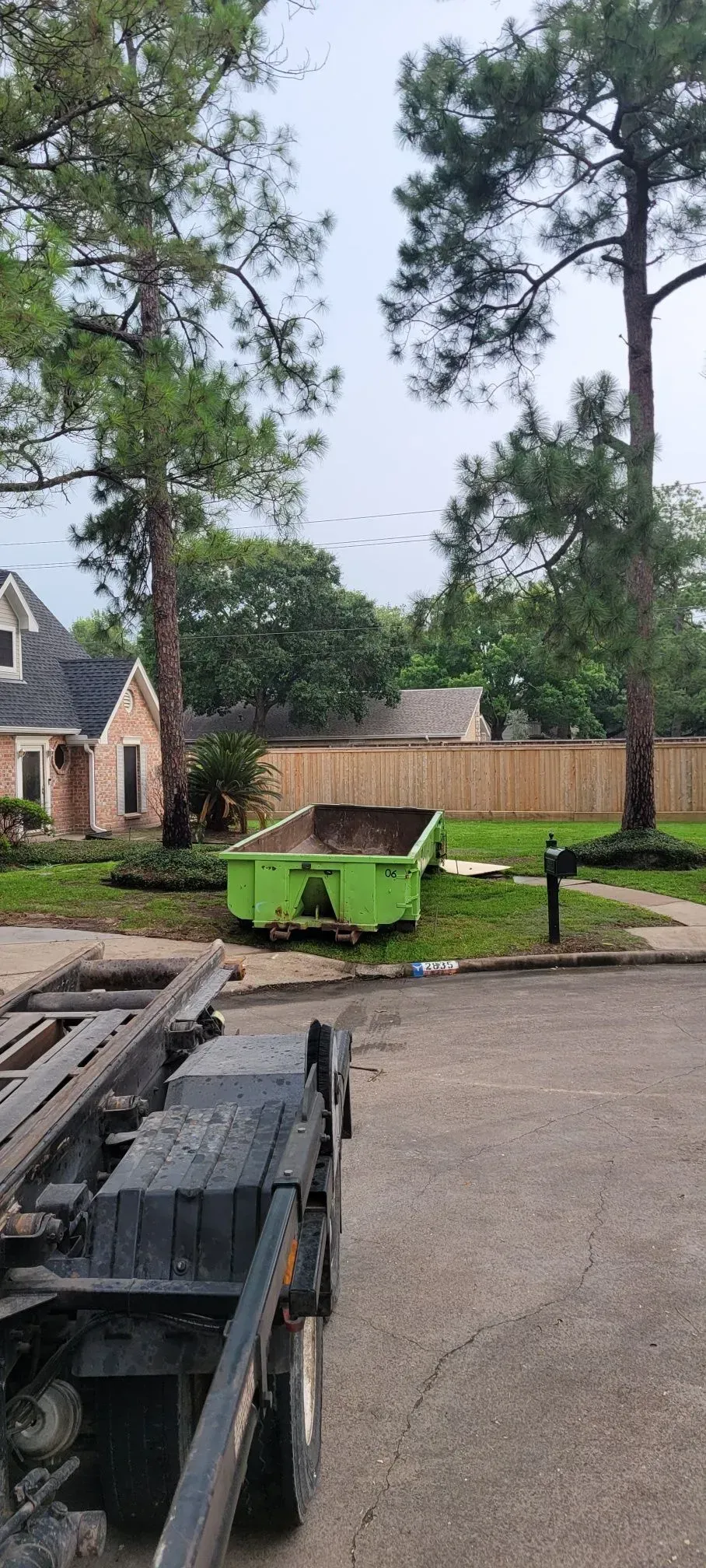 A green dumpster is in front of a house. Two tall trees and a wooden fence are in the background.