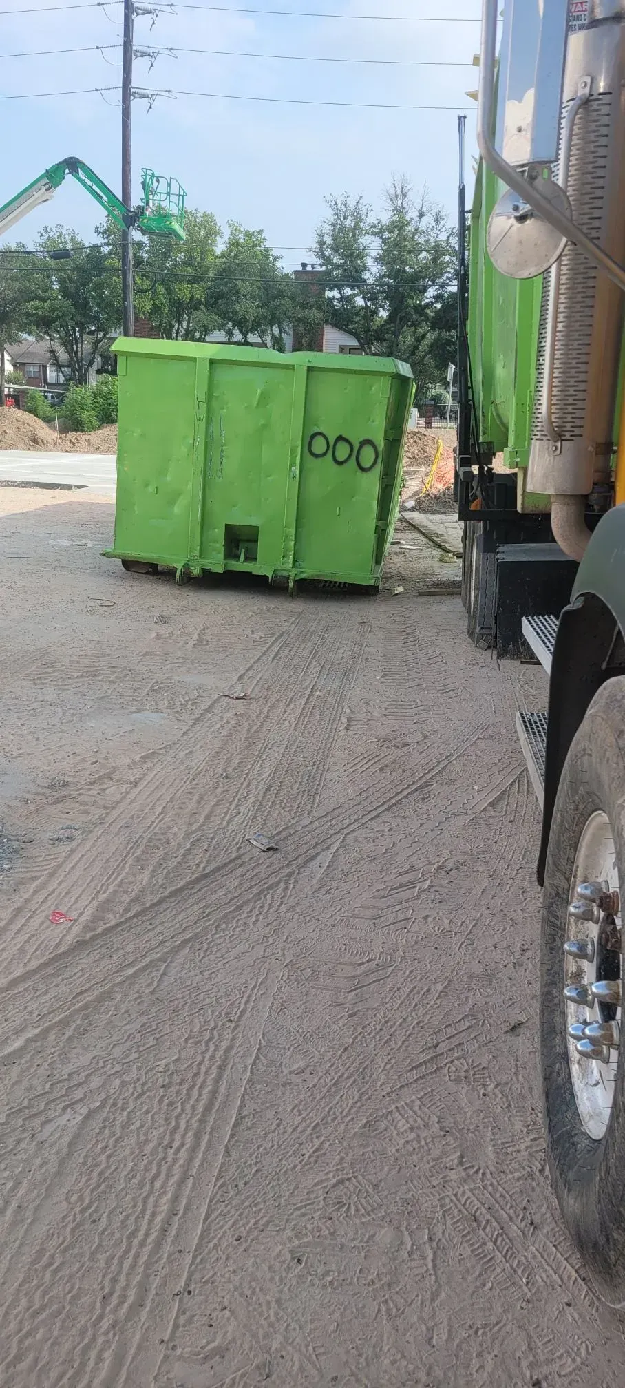 A green dumpster next to a truck, on a dirt road. Blue sky.