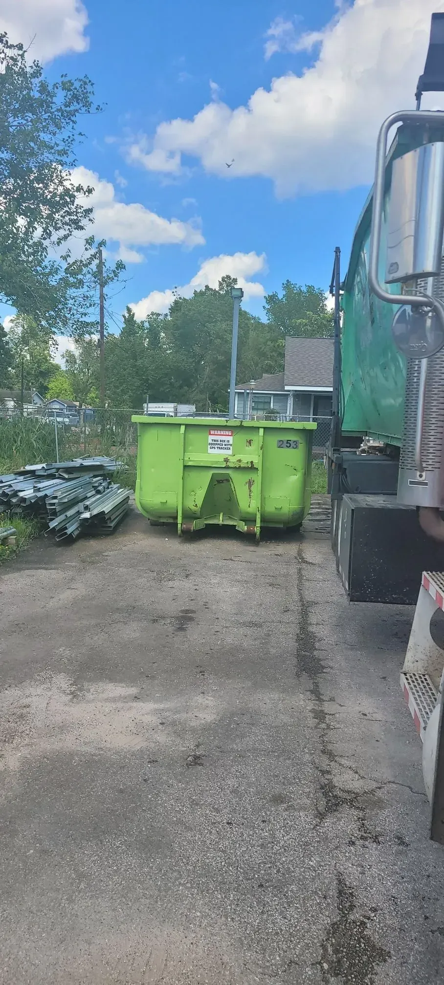 Green dumpster next to a green truck on a gravel lot under a partly cloudy sky.