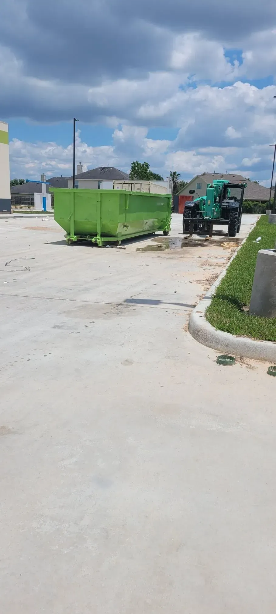 A green dumpster sits on a concrete lot near a small green tractor. Blue sky with clouds.