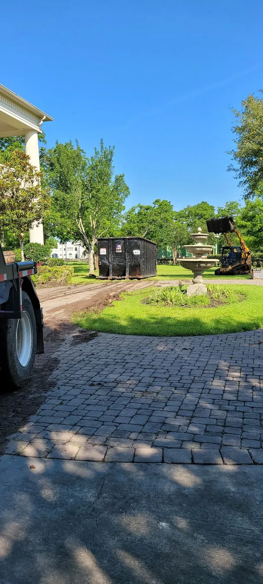 A landscaping scene with a dumpster, equipment, and a brick driveway. Bright blue sky.