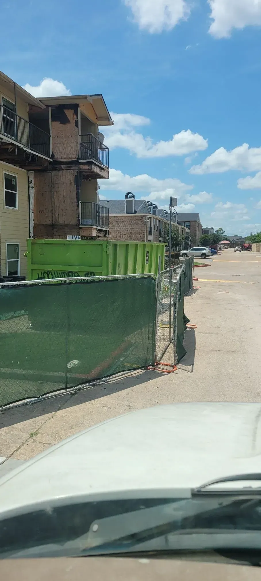 Buildings under construction behind a green fence, viewed from a car's windshield on a bright day.