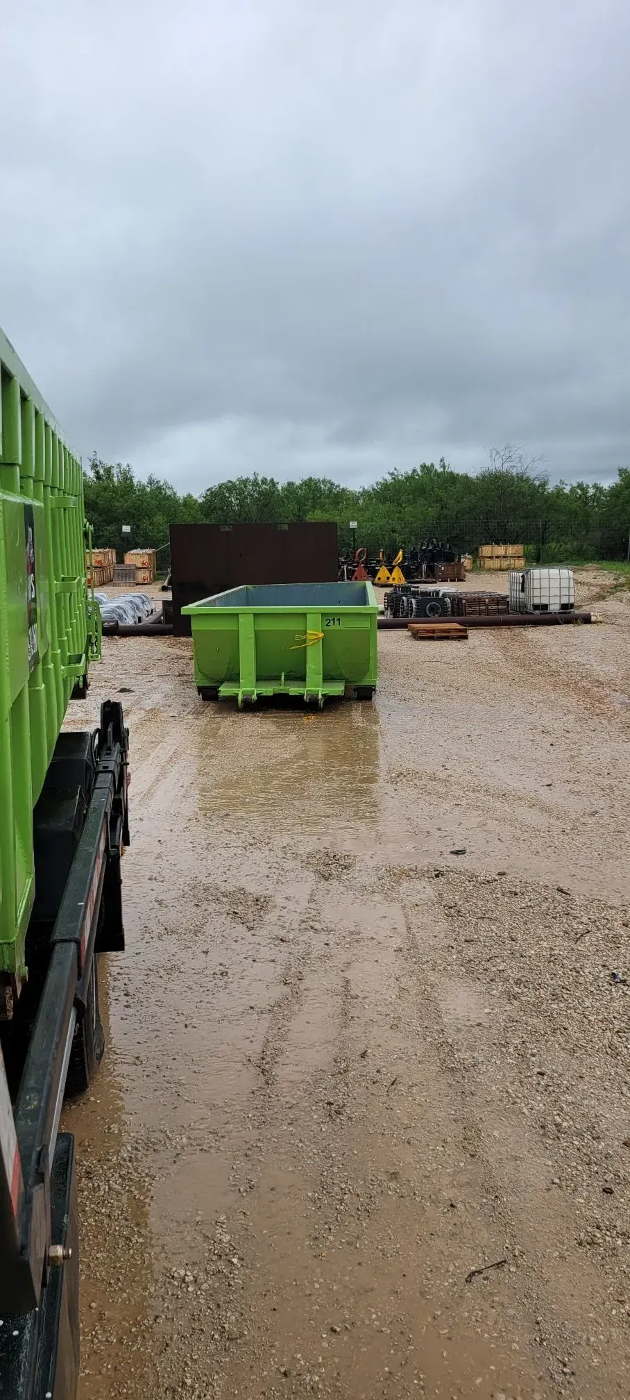 A green dumpster in a muddy lot under a cloudy sky, seen from a truck.