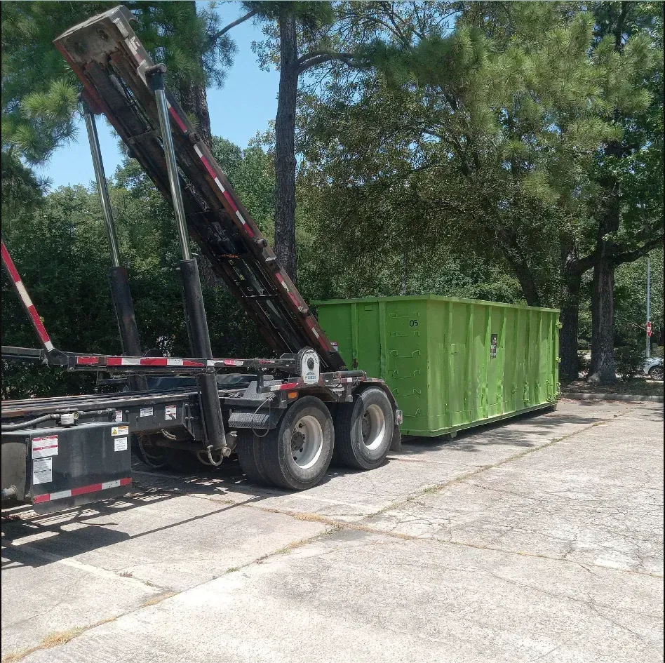 A green dumpster being unloaded from a truck on a concrete surface, surrounded by trees.