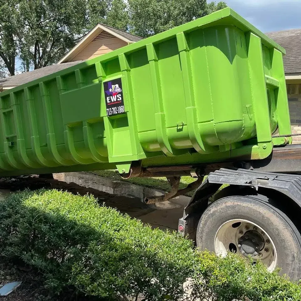 Green dumpster on a truck, parked in front of a house, above a green bush.