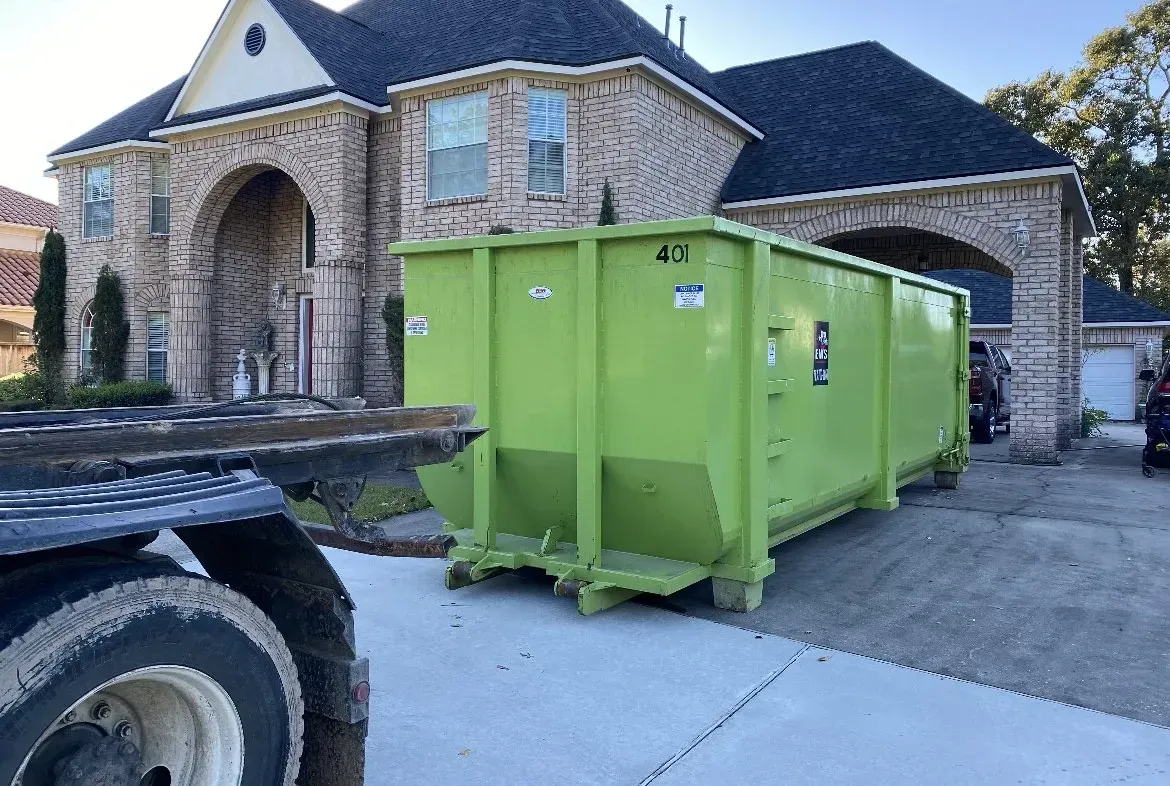 Green dumpster in a driveway next to a brick house. A truck is partially visible in the left.