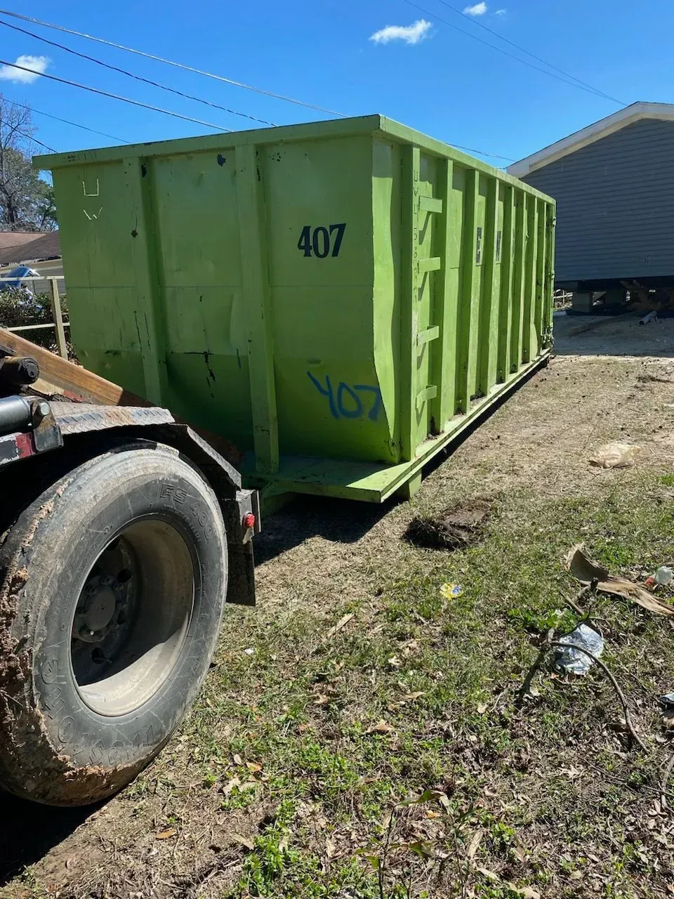 Green dumpster being towed by a truck, parked on a grassy area near a house on a sunny day.
