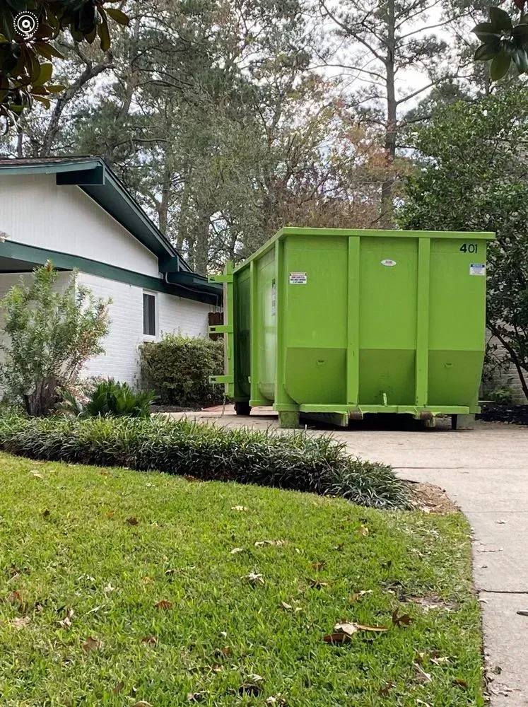 Green dumpster next to a white house with green roof and a grassy yard.