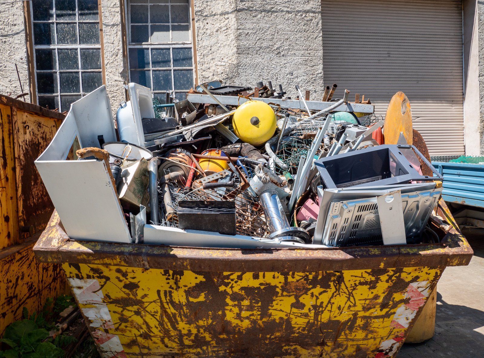 Yellow dumpster overflowing with metal scrap in front of a brick building.