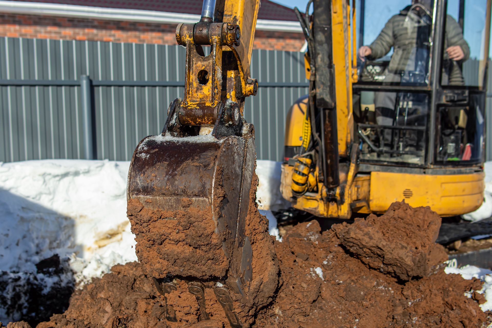 Une pelleteuse jaune soulève un godet boueux à côté d'un banc de neige sur un chantier de construction.