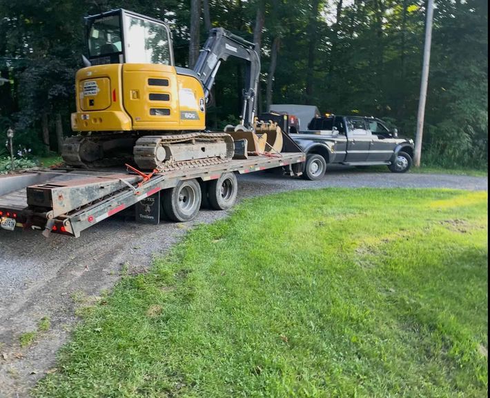 Une pelle mécanique jaune, montée sur une remorque à plateau, est tractée par une camionnette en bordure d'une route de gravier, près d'arbres.