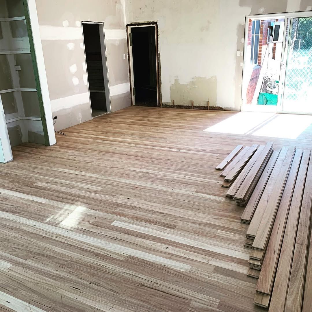 Interior room with unfinished hardwood flooring being installed. Sunlight streams in from a doorway. Drywall is visible.