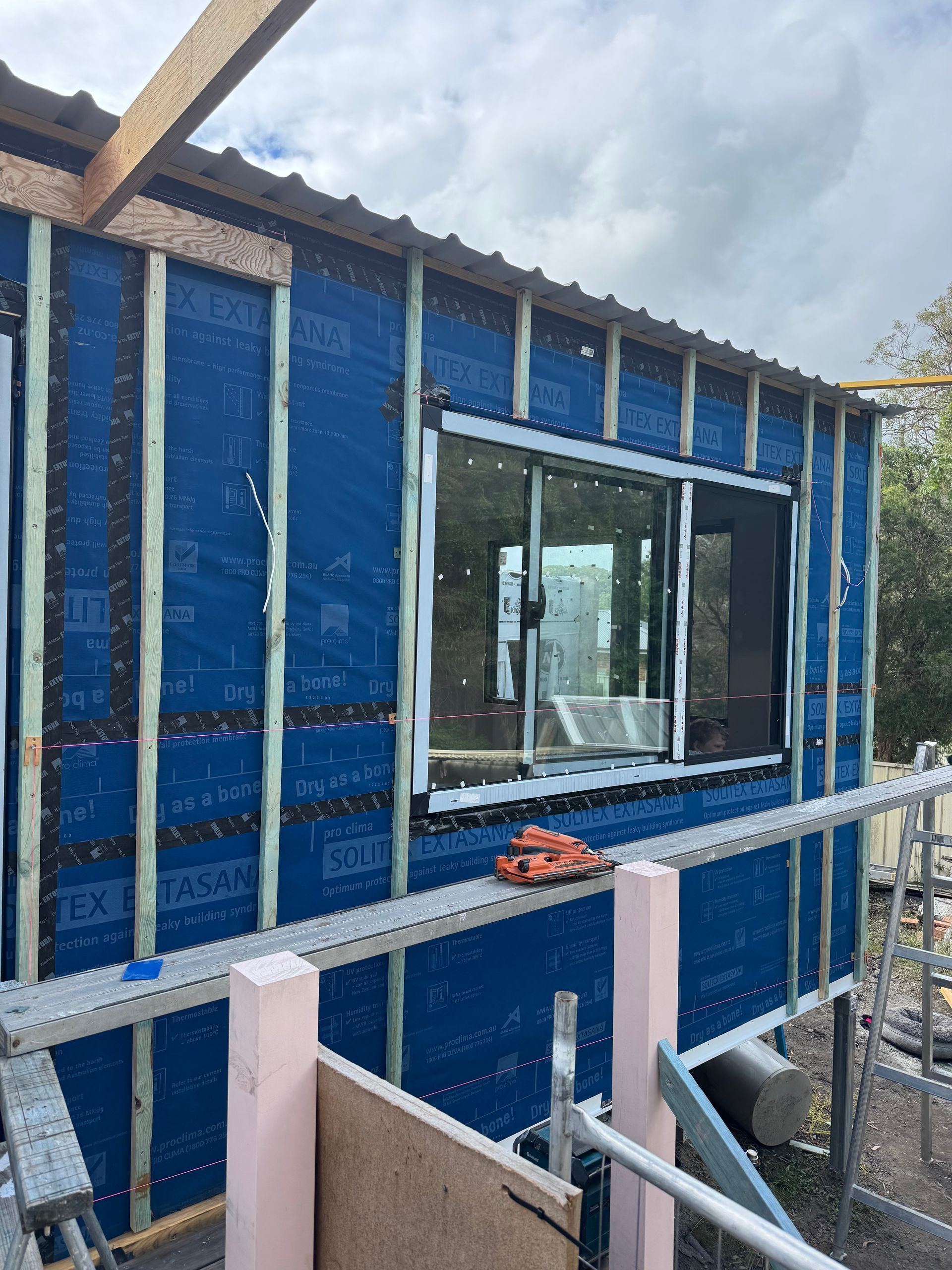 Construction site with a partially built wooden roof structure. Blue sky visible above, with a dark-walled building in the center.