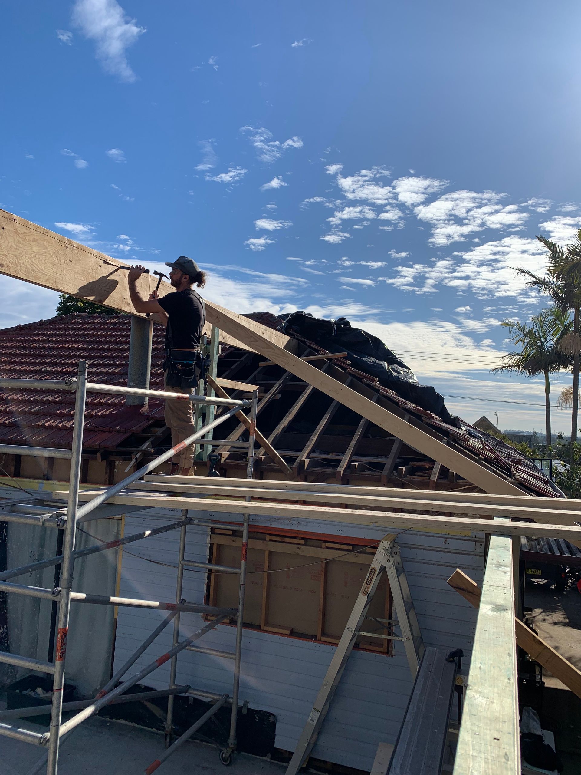 A construction worker on a rooftop, installing wood beams. Scaffolding, a blue sky, and a residential setting are visible.