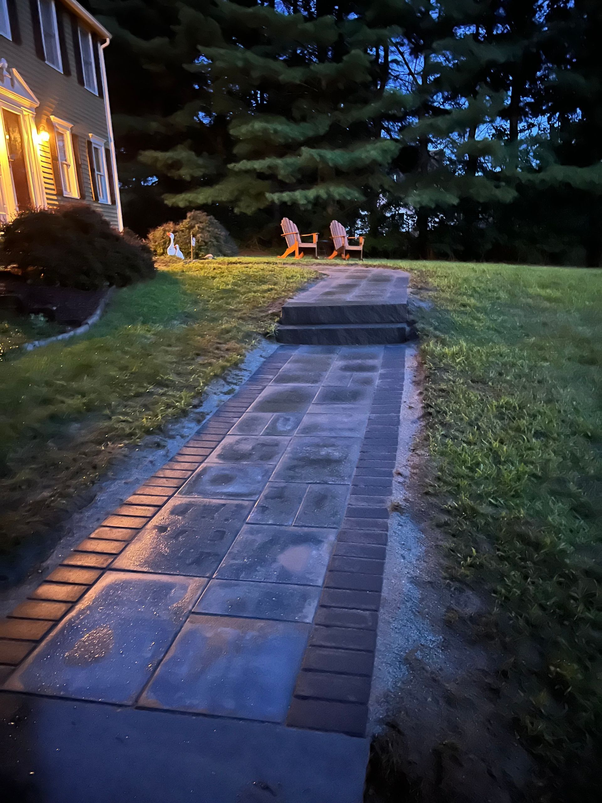 A brick walkway leading to a house with chairs on the side of it.
