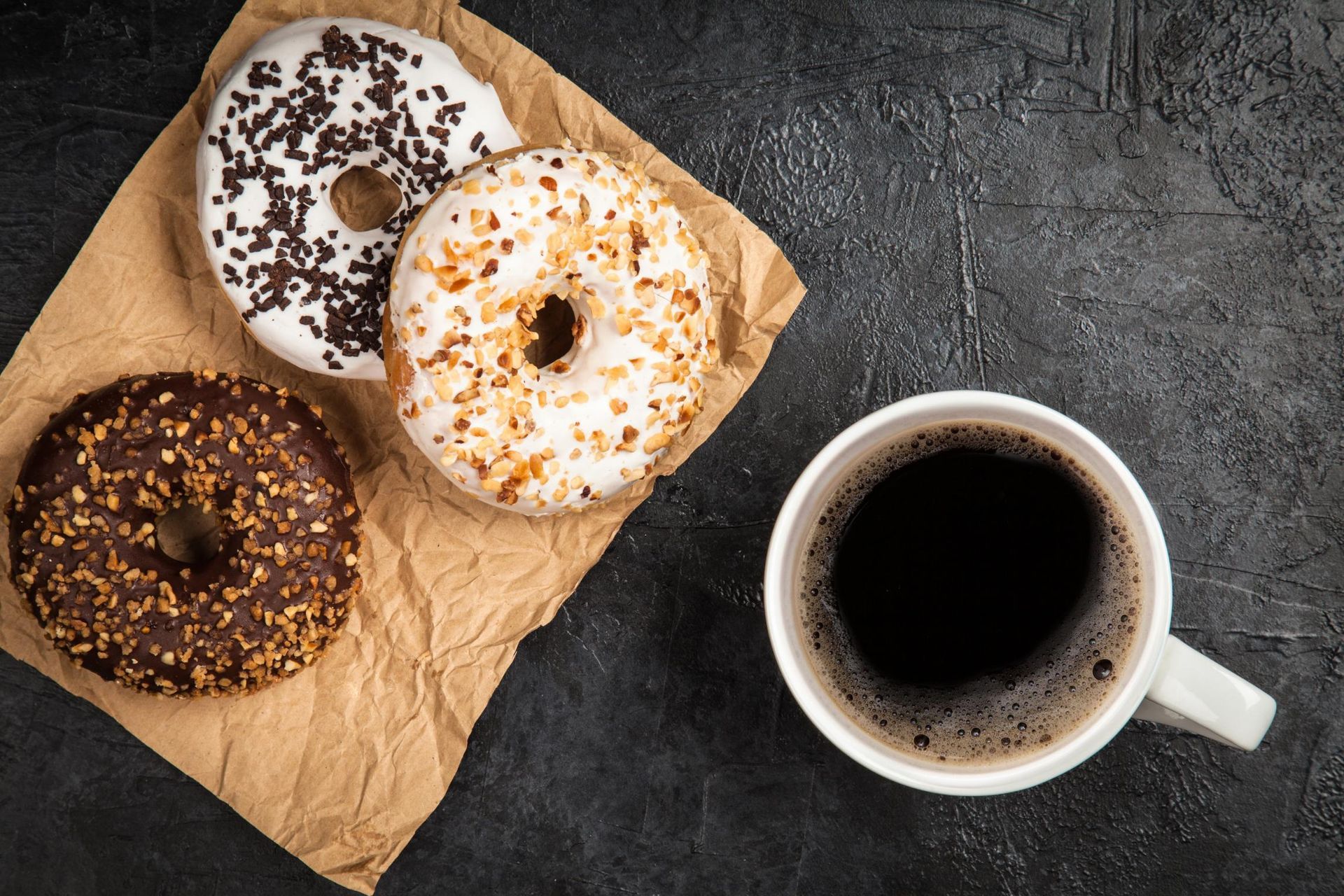 Three donuts with various toppings on a brown paper with a cup of coffee on a dark surface.