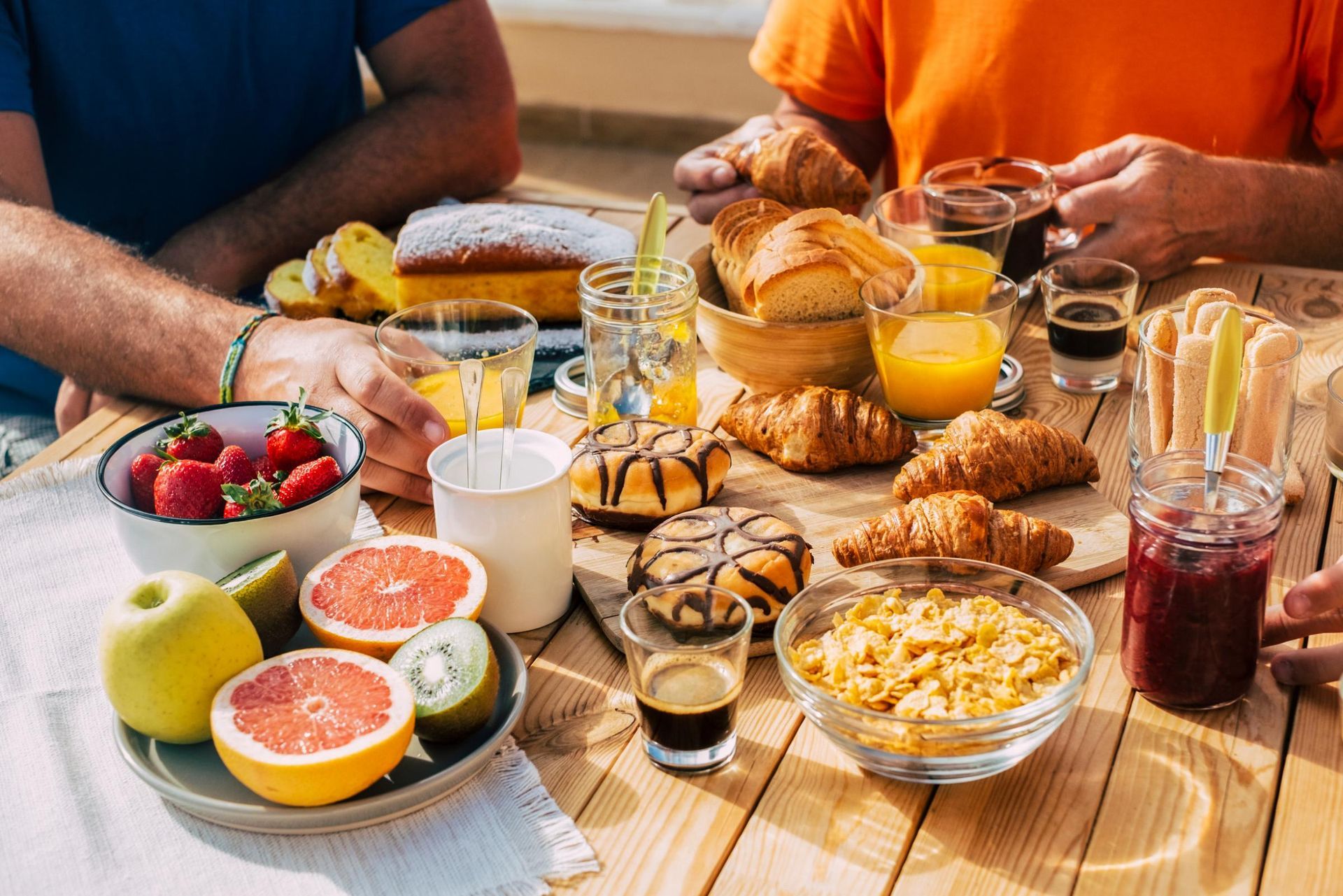 Breakfast spread on a table: fruit, pastries, juice, coffee, and people reaching for food.