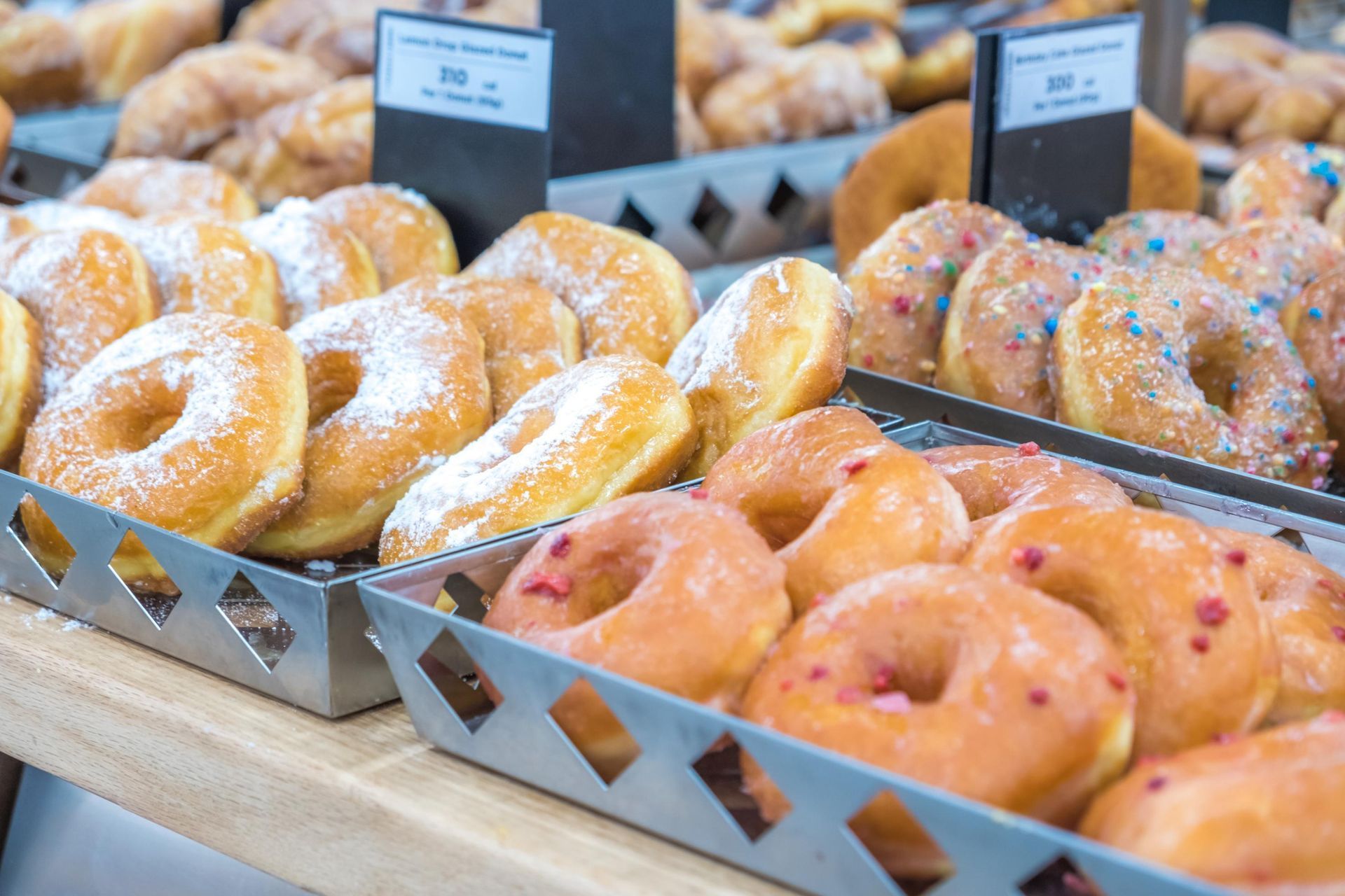 Donuts in metal trays at a bakery display; glazed, sprinkled, and powdered sugar varieties.