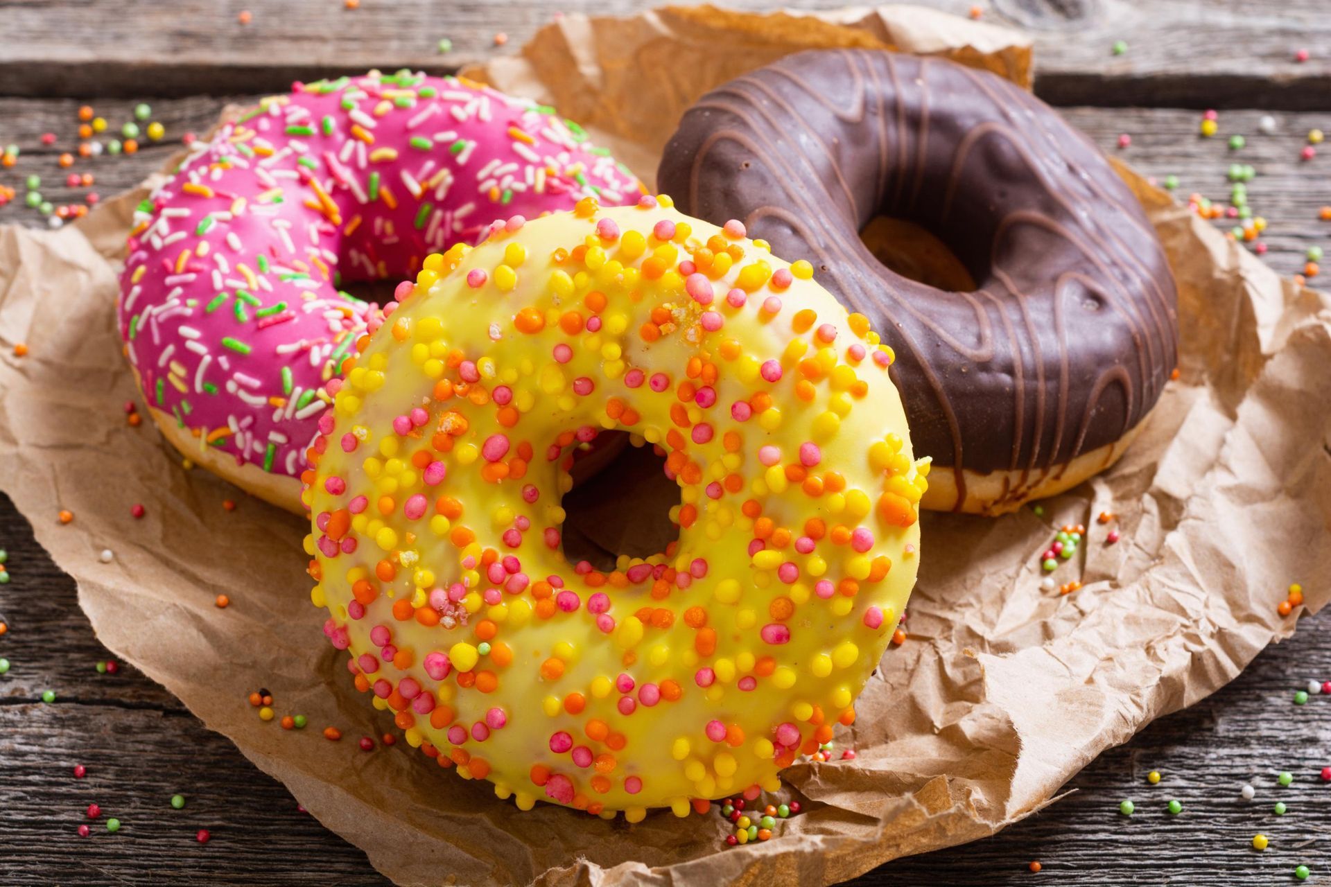 Three glazed donuts, pink, yellow, and chocolate, with sprinkles on brown paper on wood.