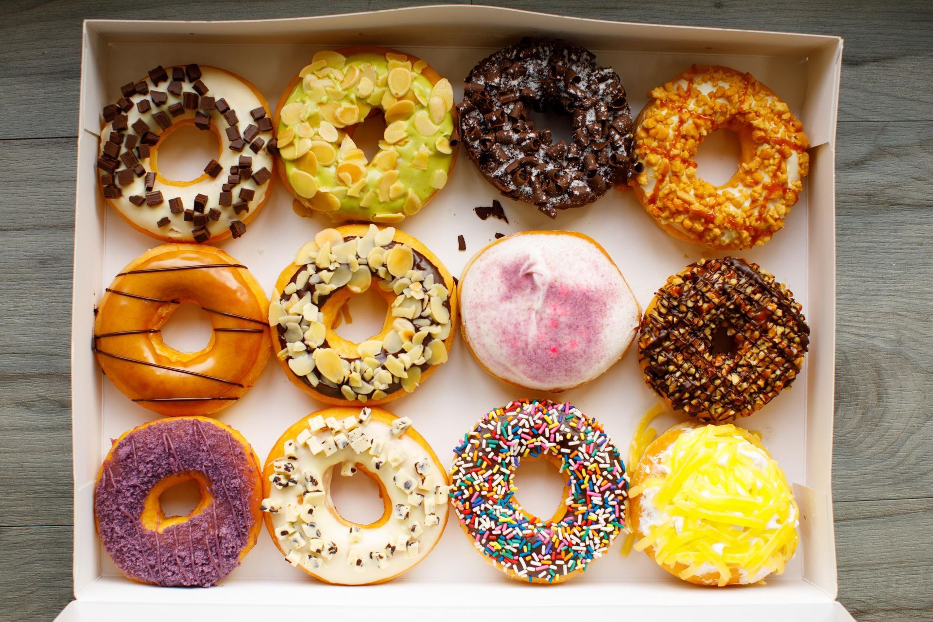 A dozen colorful donuts in a box, with various toppings and glaze.