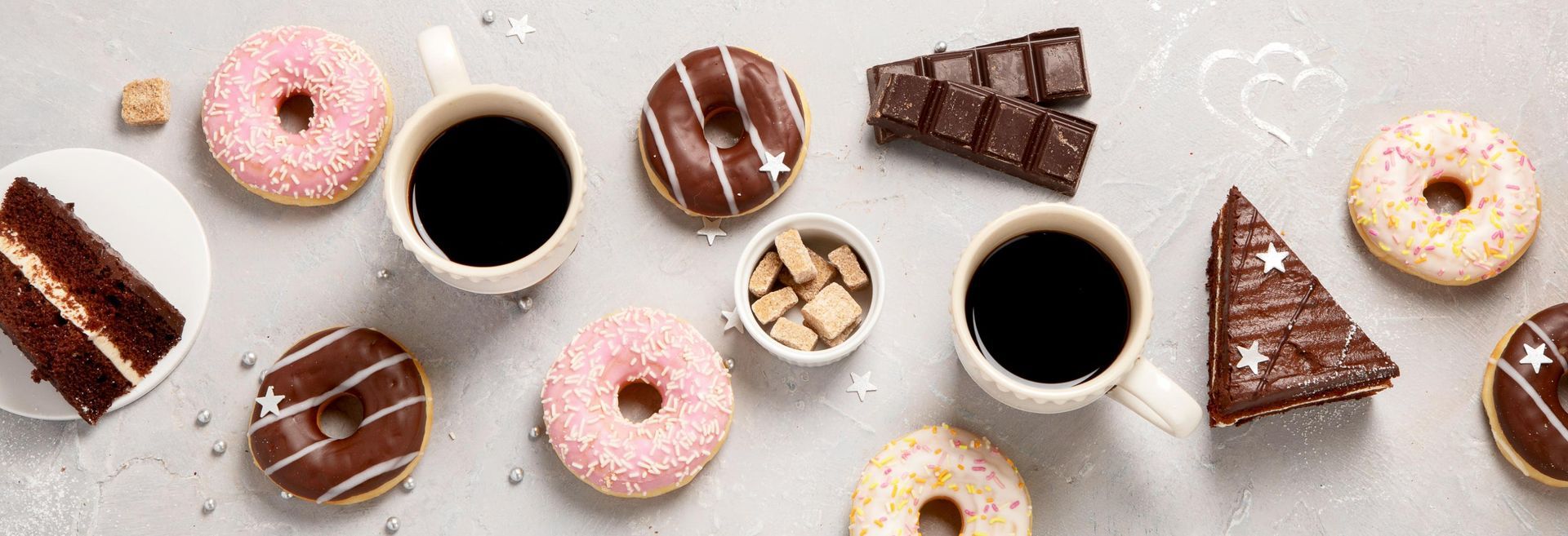 Overhead shot of donuts, coffee, chocolate, and cake on a light grey surface.