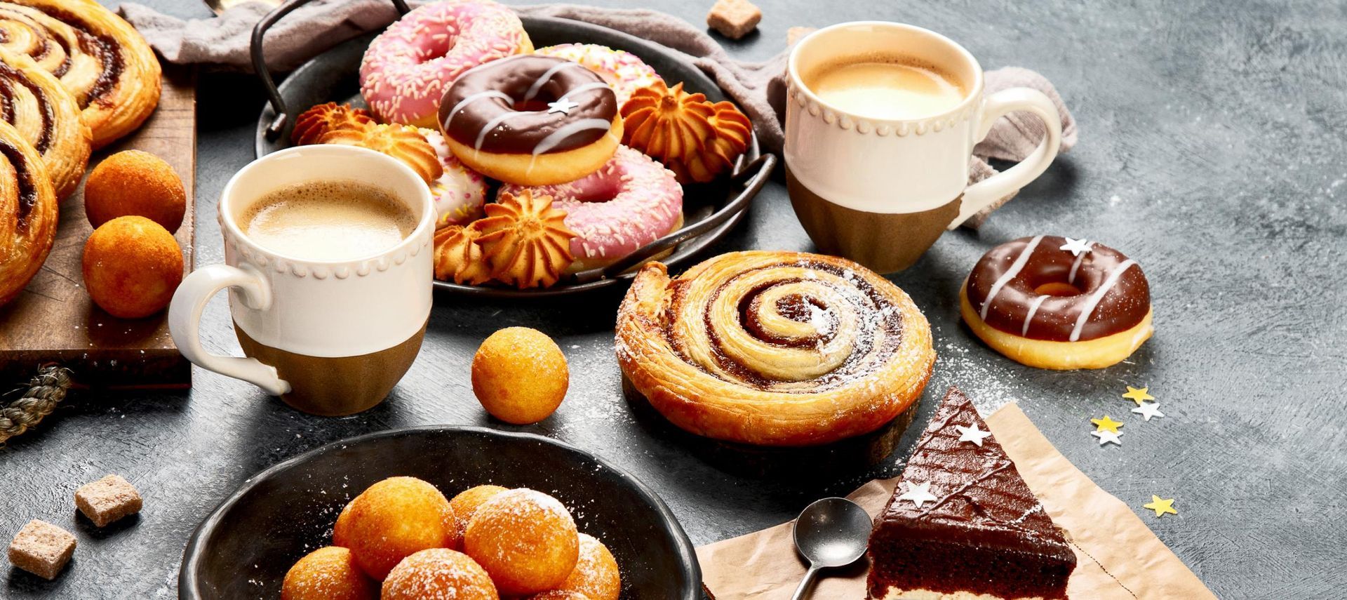 Breakfast pastries, donuts, and coffee cups on a gray surface.