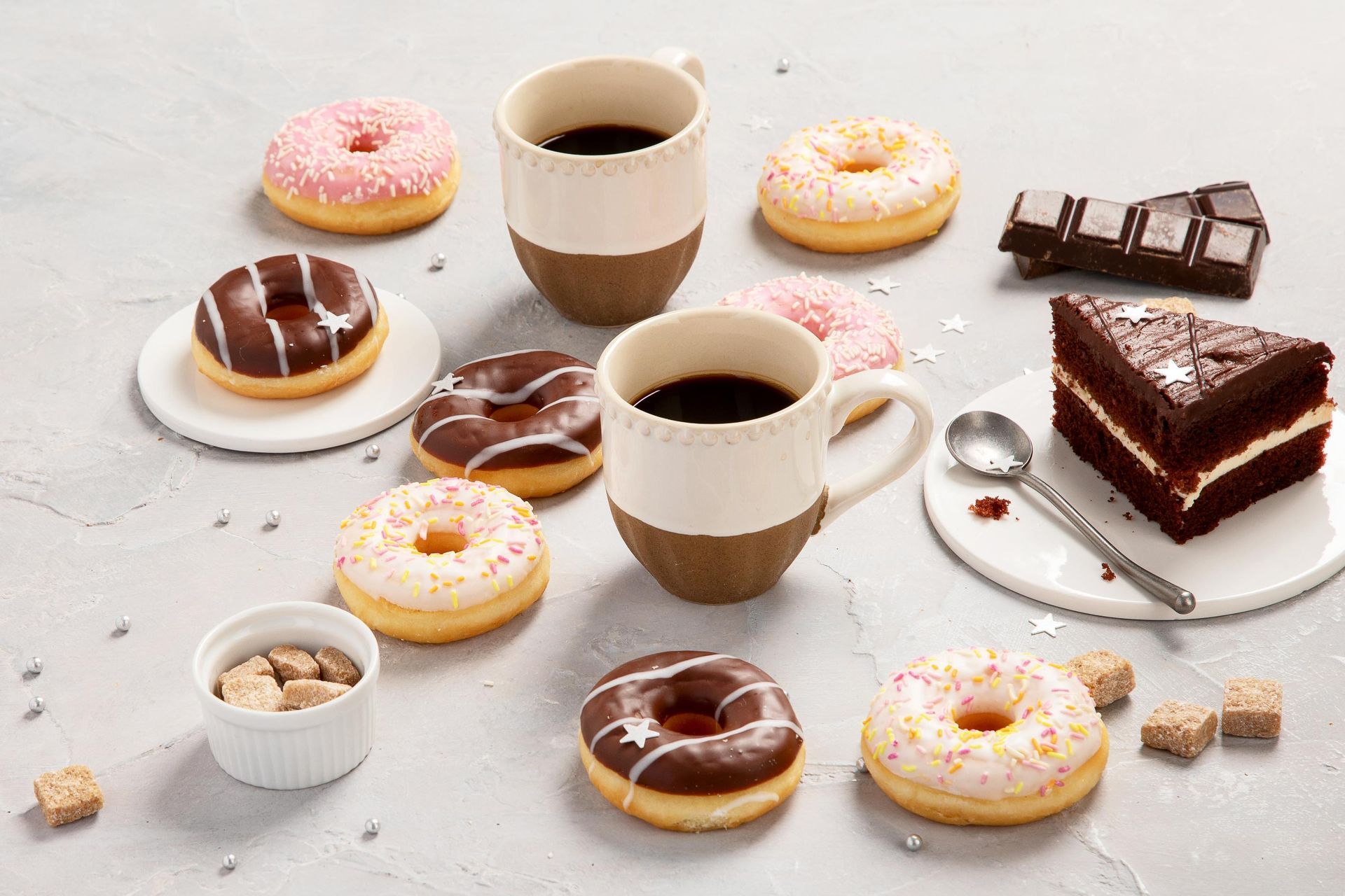 Donuts, coffee, and cake arranged on a light grey surface with sugar cubes, a spoon, and chocolate pieces.